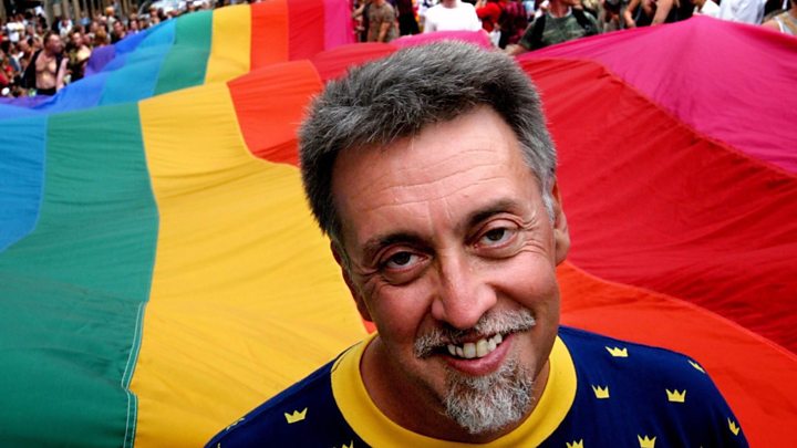Gilbert Baker, 2 de junio de 1951— 31 de marzo de 2017, con la bandera arcoiris de fondo
